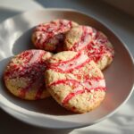 Close-up of four festive Candy Cane Shortbread Cookies with red swirls and crushed candy cane pieces on a light-colored plate.