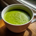 Close-up of a bowl of Creamy Pea & Mint Soup, a vibrant green soup, in a white mug on a wooden surface.