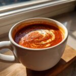 A close-up of a white mug filled with creamy tomato soup, swirled with cream and sprinkled with herbs.