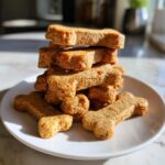 A tall stack of homemade, bone-shaped dog treats on a white plate, perfect for DIY Pet Treat Gifts for Dog Lovers.