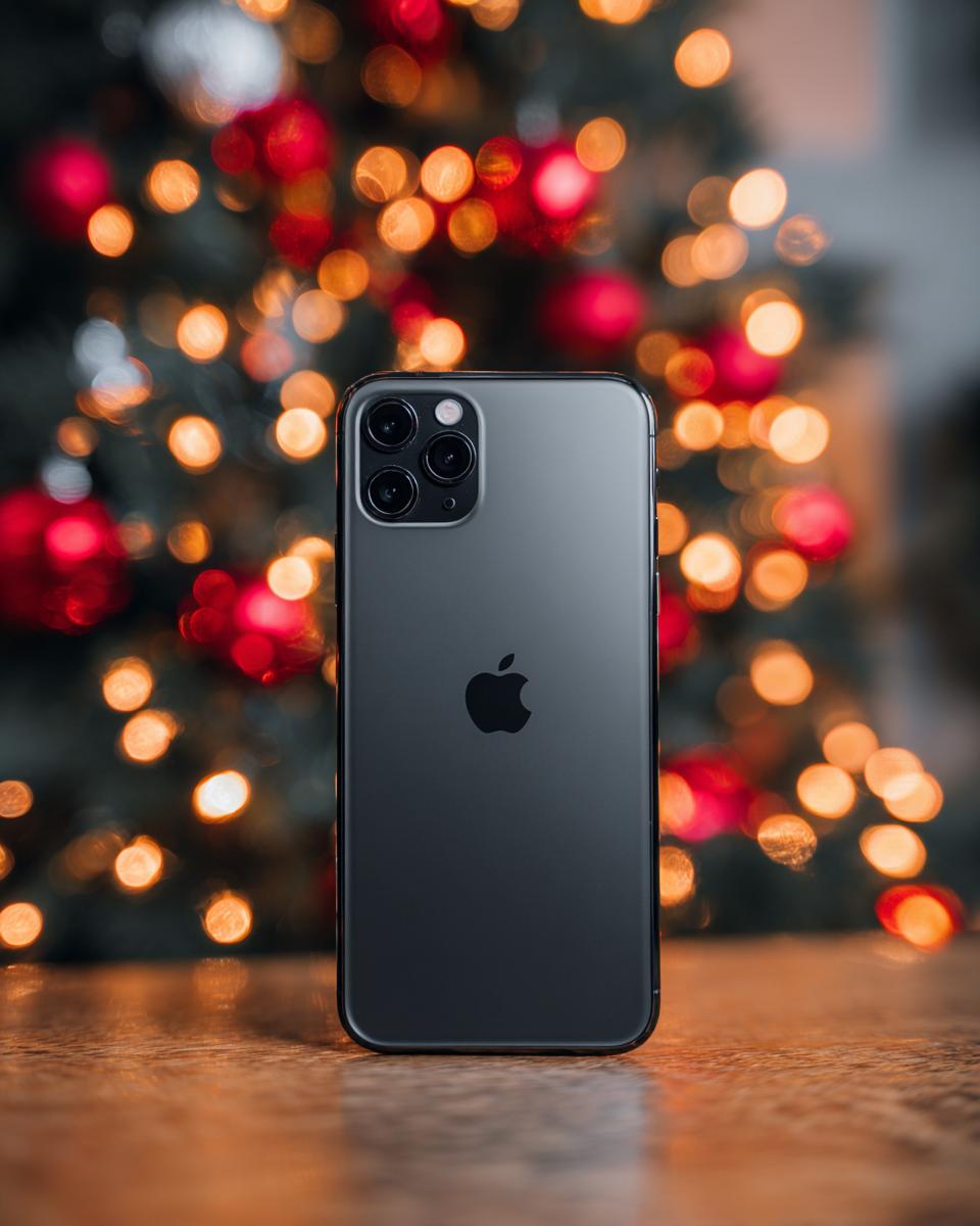 A dark gray iPhone is placed on a wooden surface in front of a blurred Christmas tree with warm, glowing lights.