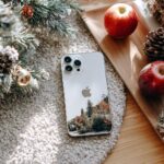 iPhone with an autumn landscape wallpaper placed on a fluffy rug next to a frosted Christmas tree branch and red apples.