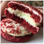 Close-up of a cut Red Velvet Cookie sandwich, revealing a thick cream cheese filling and red cookie halves dusted with crumbs.