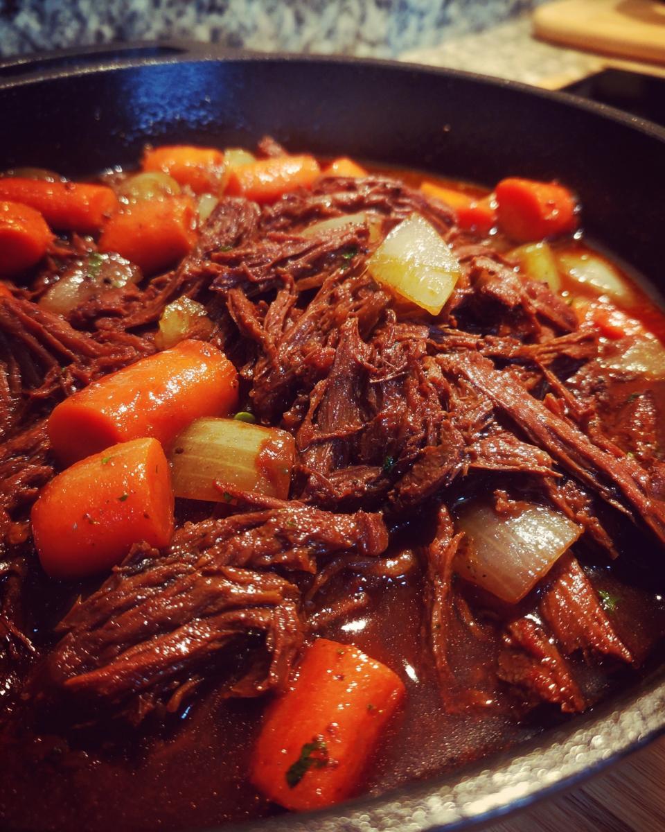 Close-up of a Rustic Red Wine Pot Roast with carrots, onions, and tender shredded beef in a cast iron skillet.