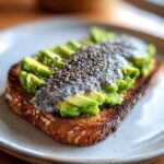 Close-up of a slice of toasted bread topped with mashed avocado and savory chia pudding, sprinkled with chia seeds.