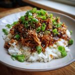 A bowl of fluffy white rice topped with shredded Slow Cooker Teriyaki Chicken, garnished with green onions and sesame seeds.