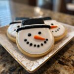 Close-up of adorable Snowman Hat Cookies decorated with white icing, black hats, and orange carrot noses.