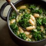 Close-up of Tuscan White Bean & Kale Soup in a black pot with a spoon scooping up the soup.