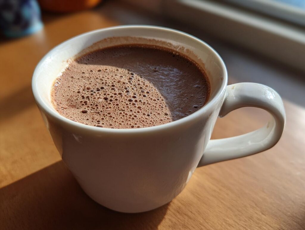 A mug of The Creamiest Chocolate Recipe, with foamy texture on top, on a wooden surface.