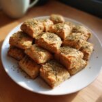 Pile of freshly baked catnip-infused cat treats on a white plate, sprinkled with herbs.