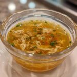 Close-up of a clear glass bowl filled with fish broth for cats, garnished with herbs.