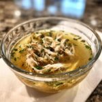 A clear glass bowl filled with fish broth for cats, topped with shredded fish and herbs. This is our Fish Broth Cat Recipe.