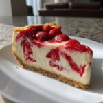 Close-up of a slice of Strawberry Shortcake Cheesecake on a white plate, showcasing the creamy filling and strawberry topping.