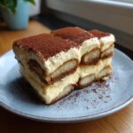 A slice of classic tiramisu on a blue plate, dusted with cocoa powder.