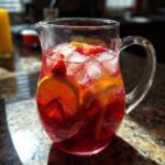 A pitcher filled with a vibrant red big batch cocktail, ice, and fruit slices, perfect for summer parties.
