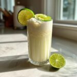 A tall glass of Coconut Lime Refresher garnished with lime slices, next to a half lime.