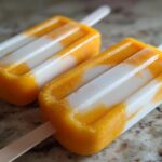 Two homemade Coconut Mango Popsicles on a countertop, showing layers of mango and coconut.