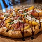 Close-up of a Grilled Peach & Prosciutto Pizza on a wooden board, with peaches, prosciutto, and balsamic glaze.