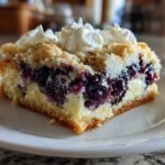 Close-up of a slice of Lemon Blueberry Cheesecake Dump Cake on a white plate, topped with whipped cream.