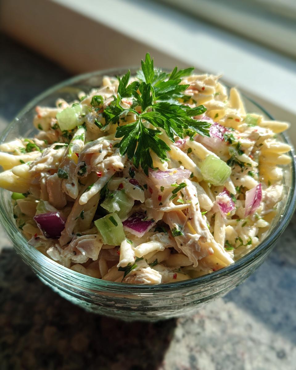 Close-up of pasta salad and chicken in a glass bowl, with red onion, celery, and herbs. The pasta salad and chicken is the primary keyword.