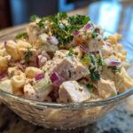 Close-up of pasta salad and chicken in a glass bowl, with red onion and fresh parsley.