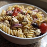 Close-up of a high protein pasta salad with chicken, tomatoes, red onion, and feta cheese. The pasta salad high protein is in a white bowl.